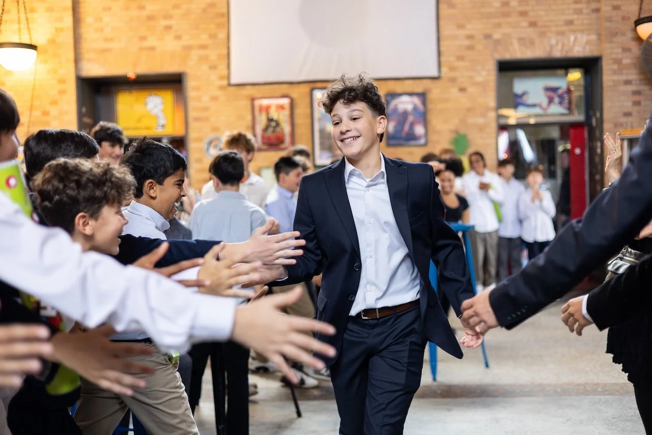 A young boy in a suit walking through a line of children reaching out to high-five him inside a school or community center with brick walls and artwork