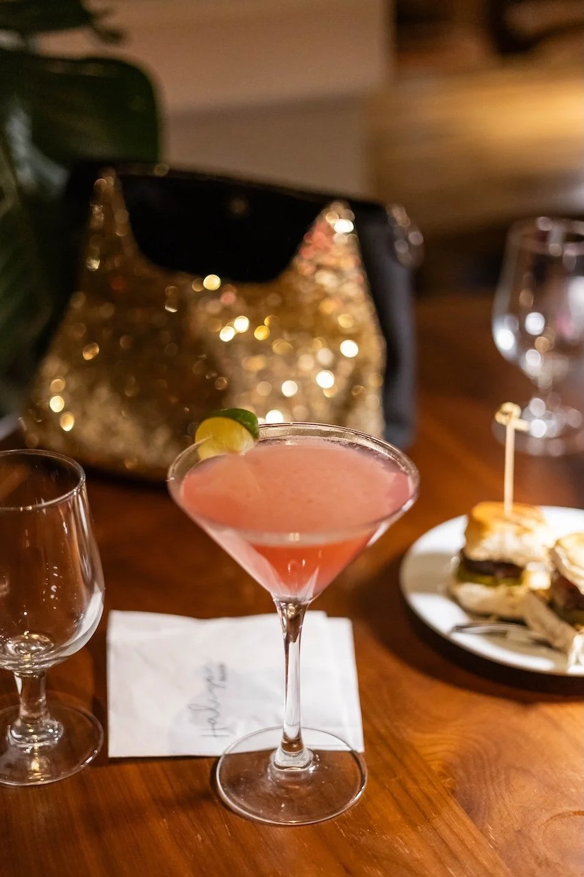 A pink cocktail with a lime wedge garnish on a wooden table at Halcyon restaurant in Montclair, NJ alongside empty wine glasses and a plate of sliders in the background.