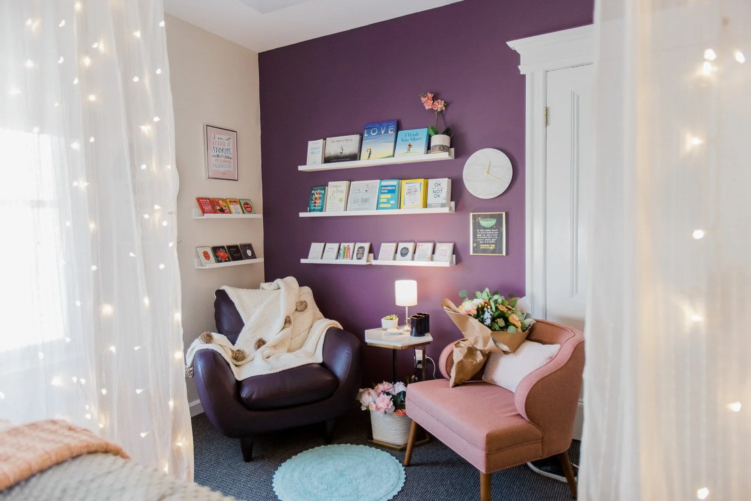 A cozy sitting area with a purple accent wall, white shelves, a pink upholstered loveseat with a bouquet of flowers on it, a dark armchair with a cream throw blanket, a small side table with a lamp, and string lights framing the view.