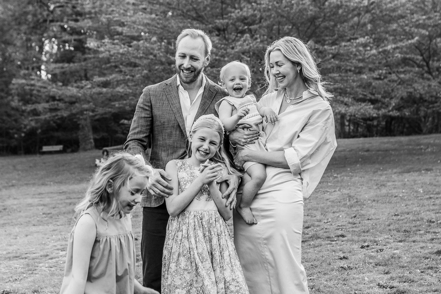 A black and white photo of a happy family of five outdoors in a park, including two adult women, a man, and three young girls, all smiling and interacting playfully.