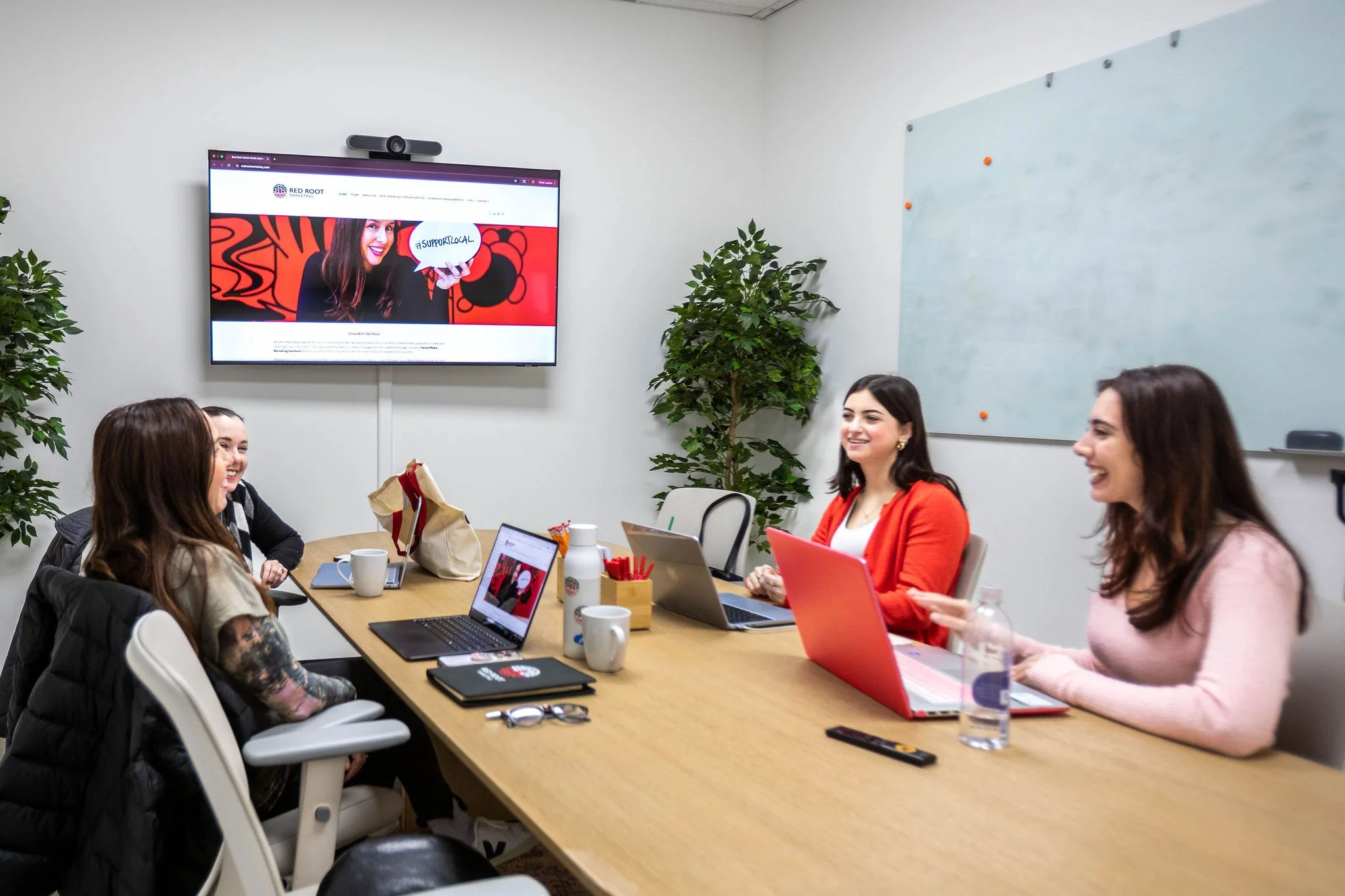 A group of five women sitting around a conference table in a meeting room, smiling and engaging in a discussion. There are laptops, notebooks, coffee cups, and water bottles on the table. A large screen on the wall displays a website with a woman hol