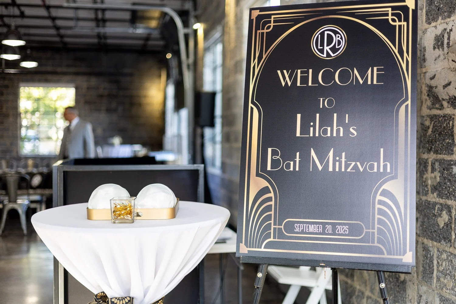 A sign welcoming guests to Liah's Bat Mitzvah on September 20, 2025, standing beside a small round table with white cloth and decorative gold and white items inside a glass container.