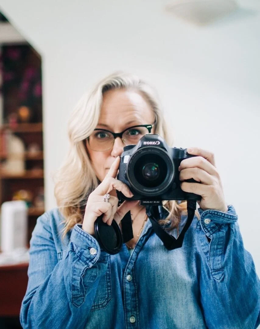 A woman with blonde hair, glasses, and a denim shirt taking a selfie with a Canon camera in an indoor setting.