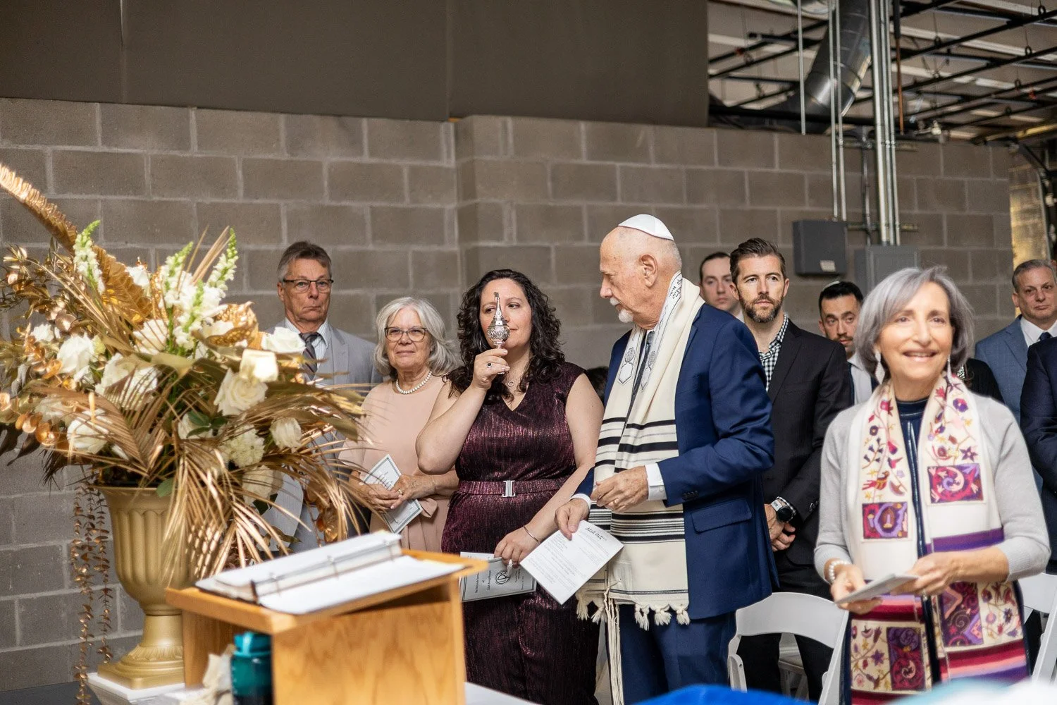 A group of diverse people praying during a religious ceremony in an urban indoor setting, with a woman smelling a dilated glass container held by a man with a kippah, beside a large flower arrangement.