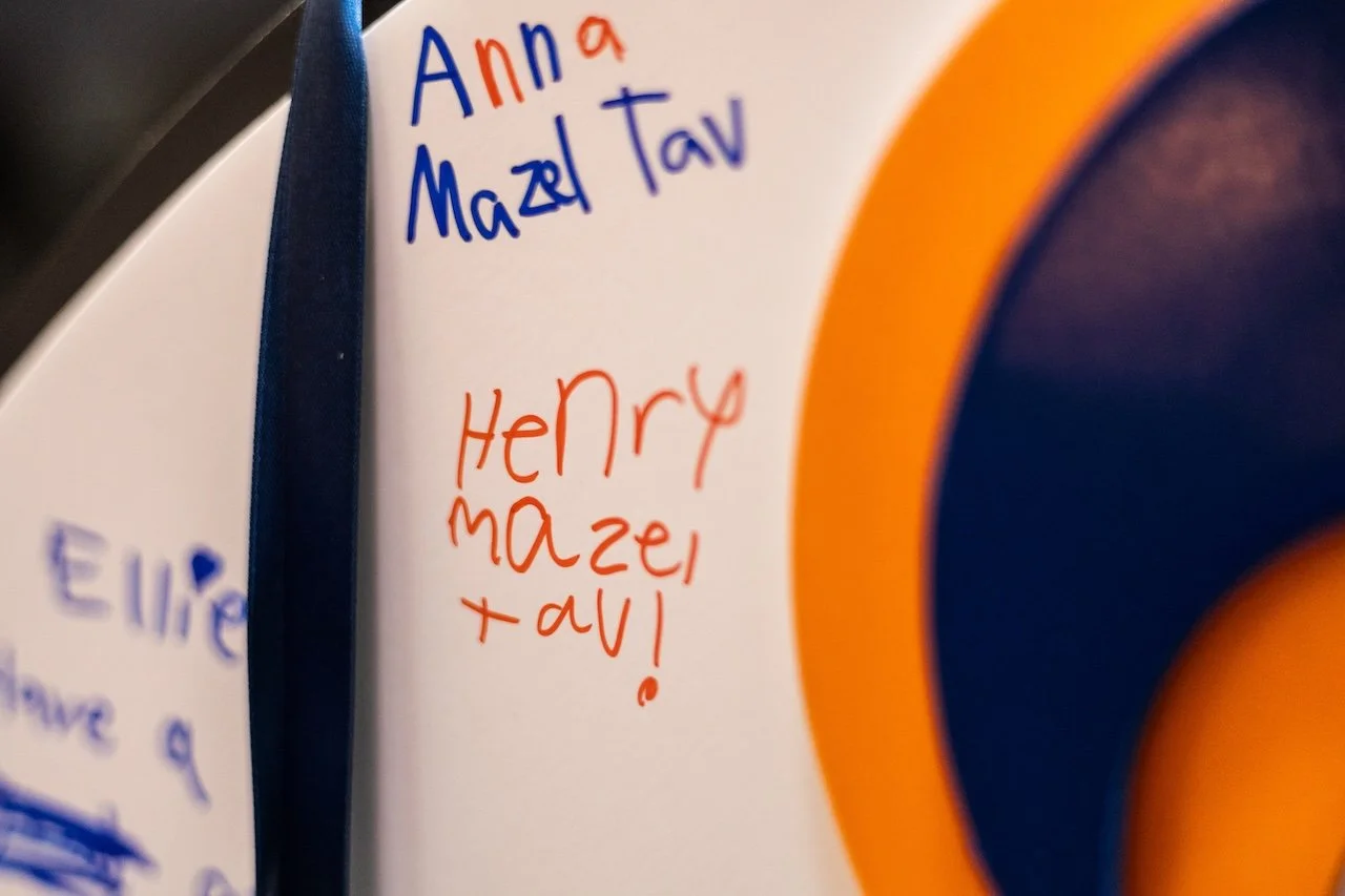 Close-up of a white surface with handwritten names 'Anna', 'Mazel Tav', and 'Henry Mazel Tavi' in blue and red marker, with a partial view of a circular orange and blue pattern.