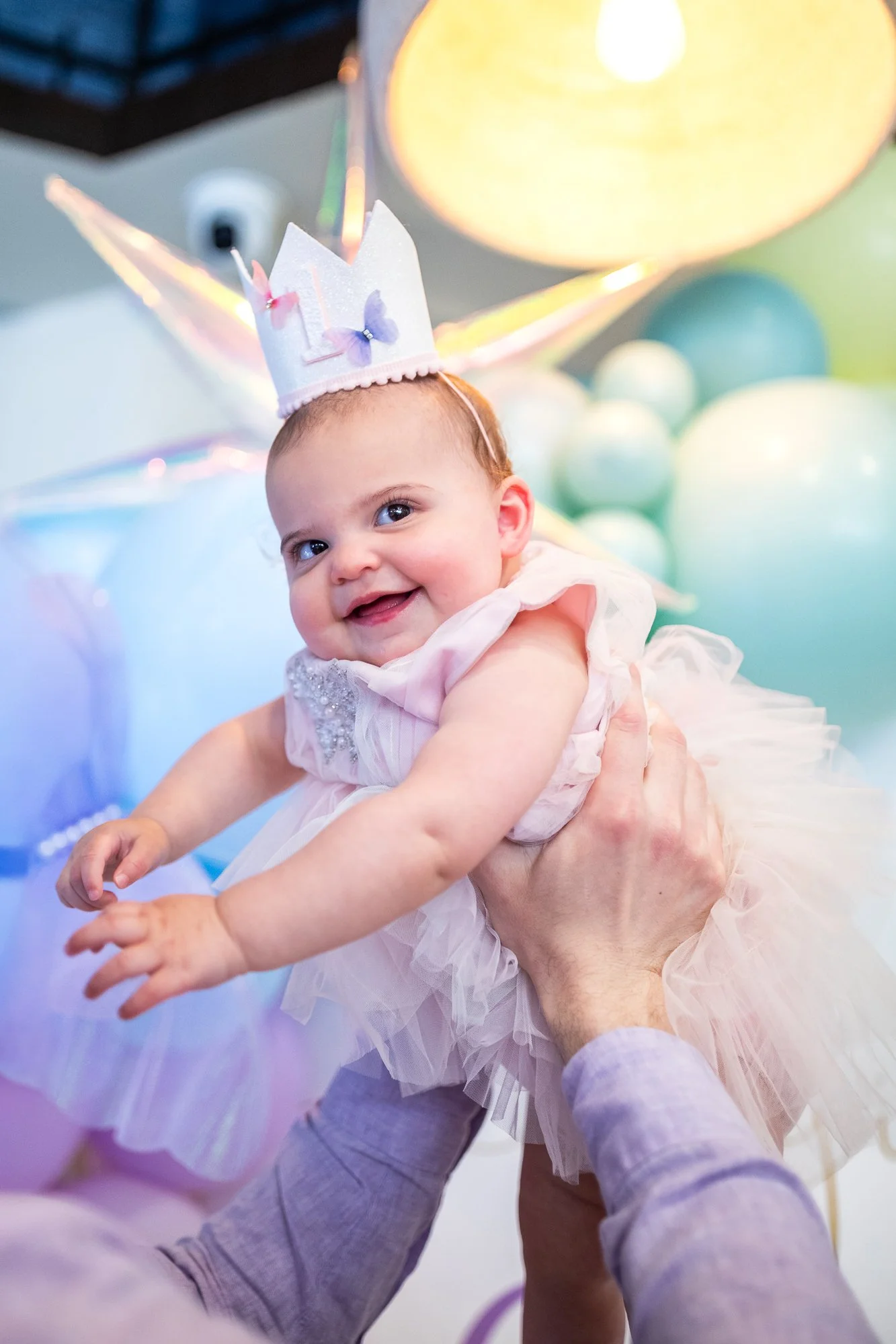 A smiling Baby girl wearing a princess crown dress being held up by adults at a celebration with pastel-colored balloons and a large yellow balloon in the background.