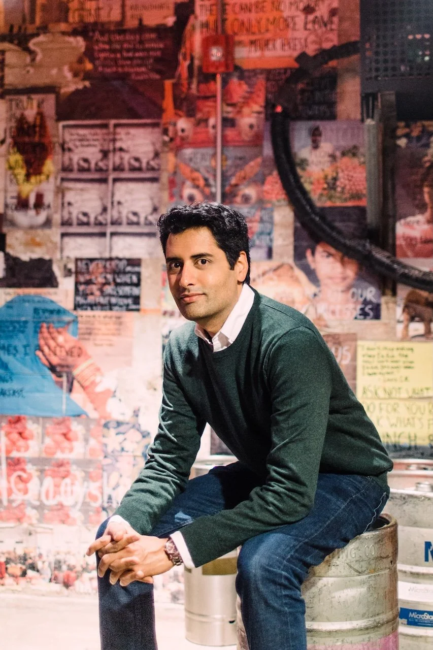 A man in a green sweater and jeans sitting on a metallic barrel against a colorful wall covered with posters and graffiti.