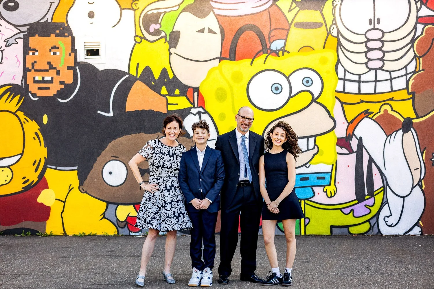 A family of four people, dressed in semi-formal attire, standing outdoor in front of a colorful mural featuring cartoon characters including SpongeBob SquarePants, Homer Simpson, Fred Flintstone, and others.