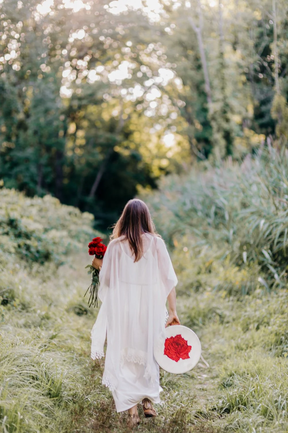 A woman dressed in white walking on a grassy path through lush greenery, holding a bouquet of red roses in her left hand and a white hat with a red flower design in her right hand, with trees and sunlight in the background.