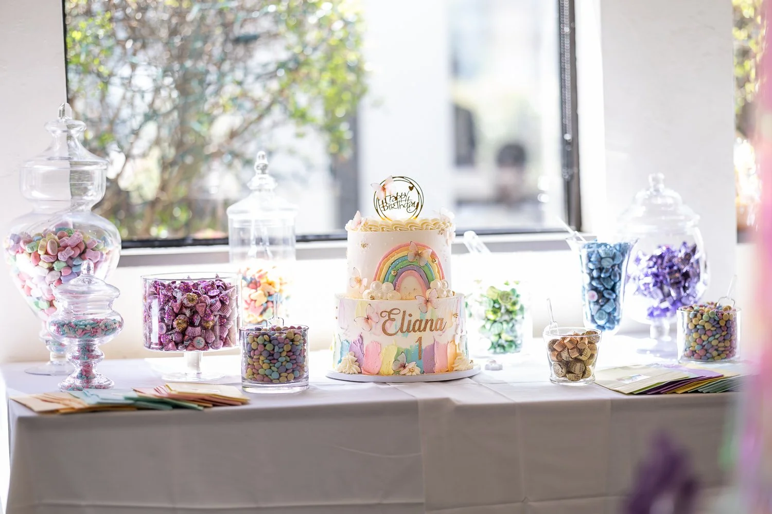 A birthday cake decorated with pastel colors, rainbows, and butterflies, with the name 'Eliana' and the number '1' on it, surrounded by jars of colorful candies on a table near a window. Candy Bar buffet by the birthday cake.
