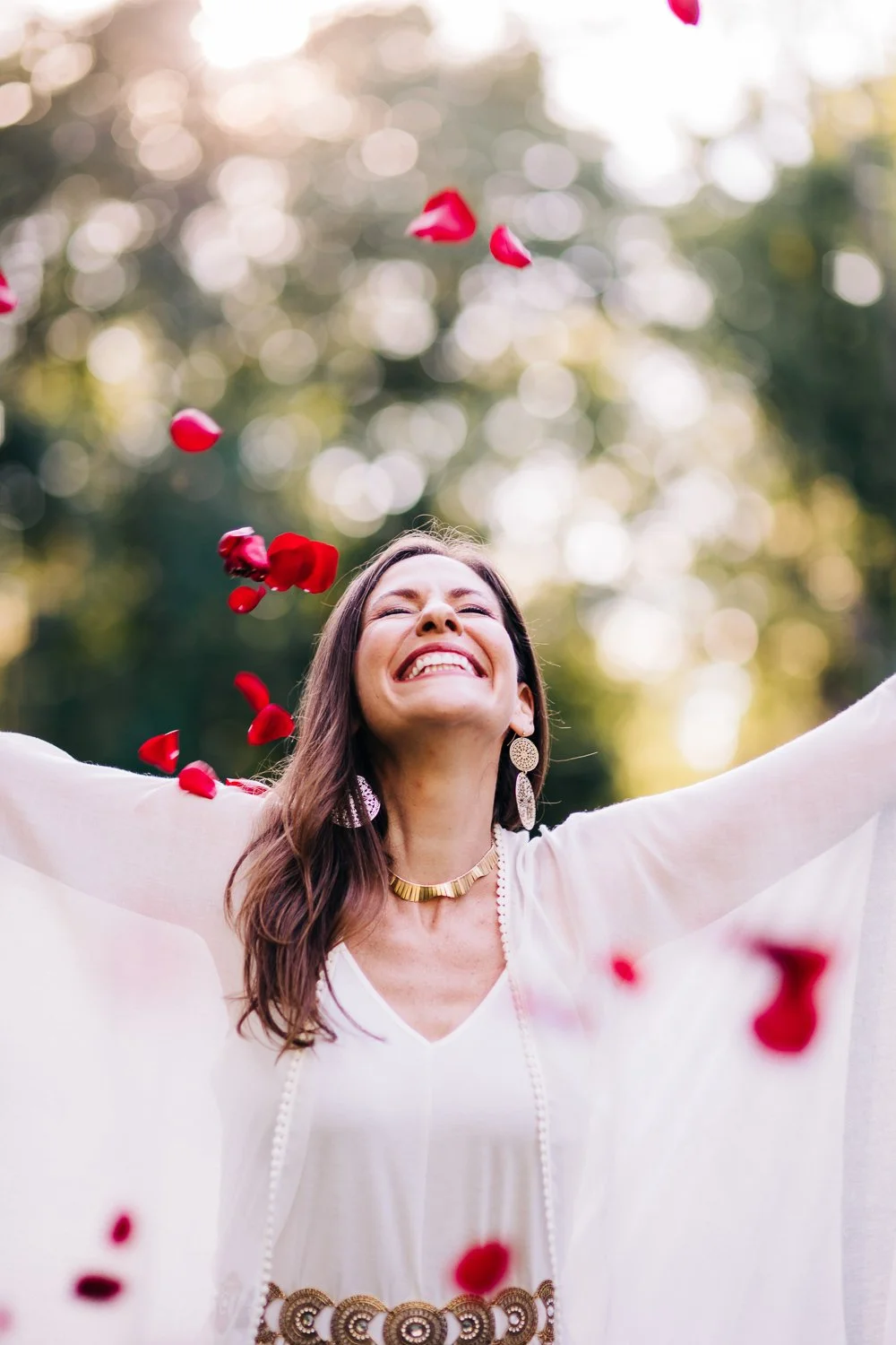 A woman outdoors smiling with her arms outstretched as red rose petals fall around her on a sunny day.