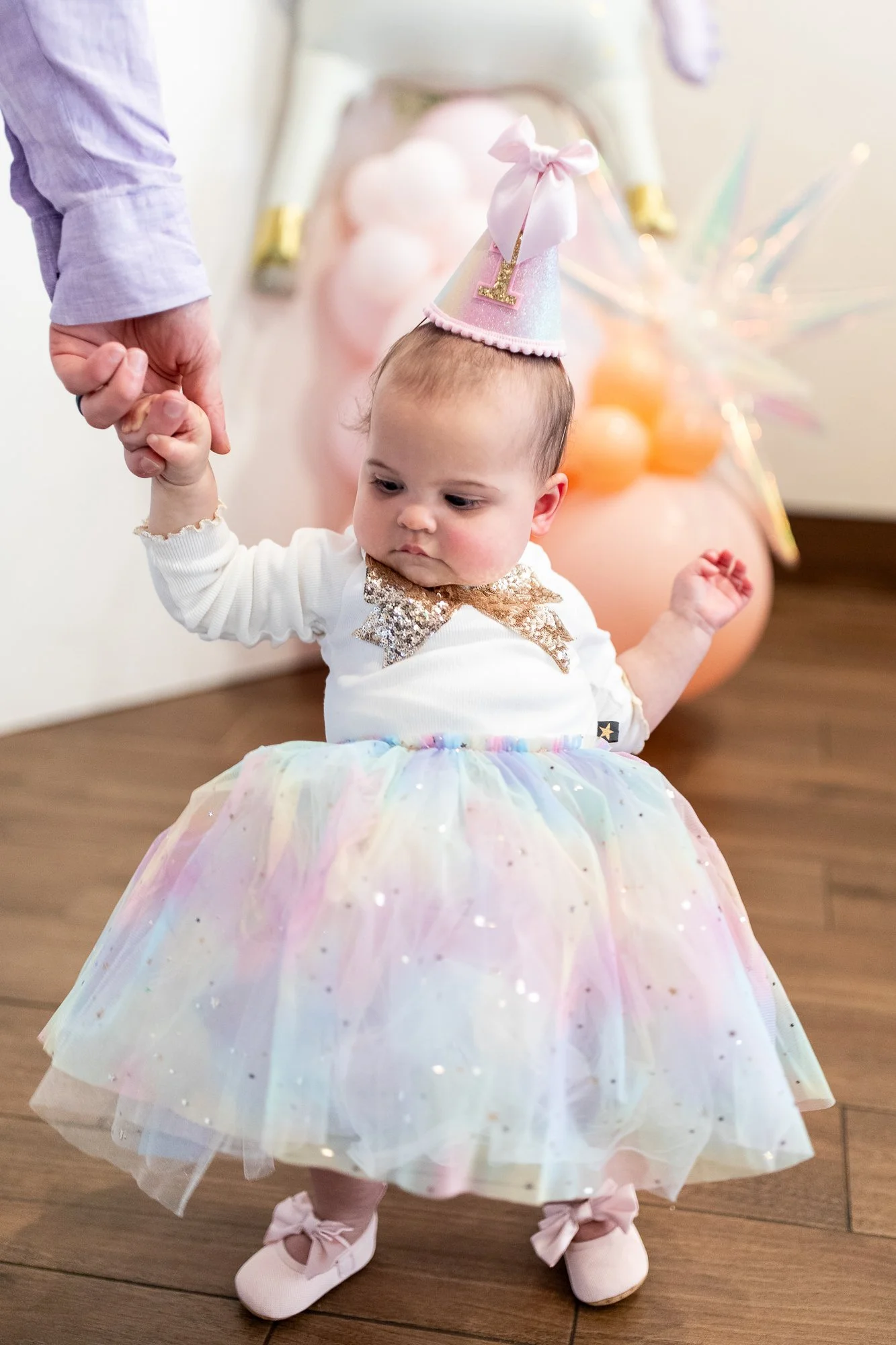A young girl dressed in a rainbow-colored tutu dress and pink ballet shoes, wearing a pink birthday hat with a letter 'L' on it, holding hands with an adult. The background features pink and orange balloons and colorful decorations.