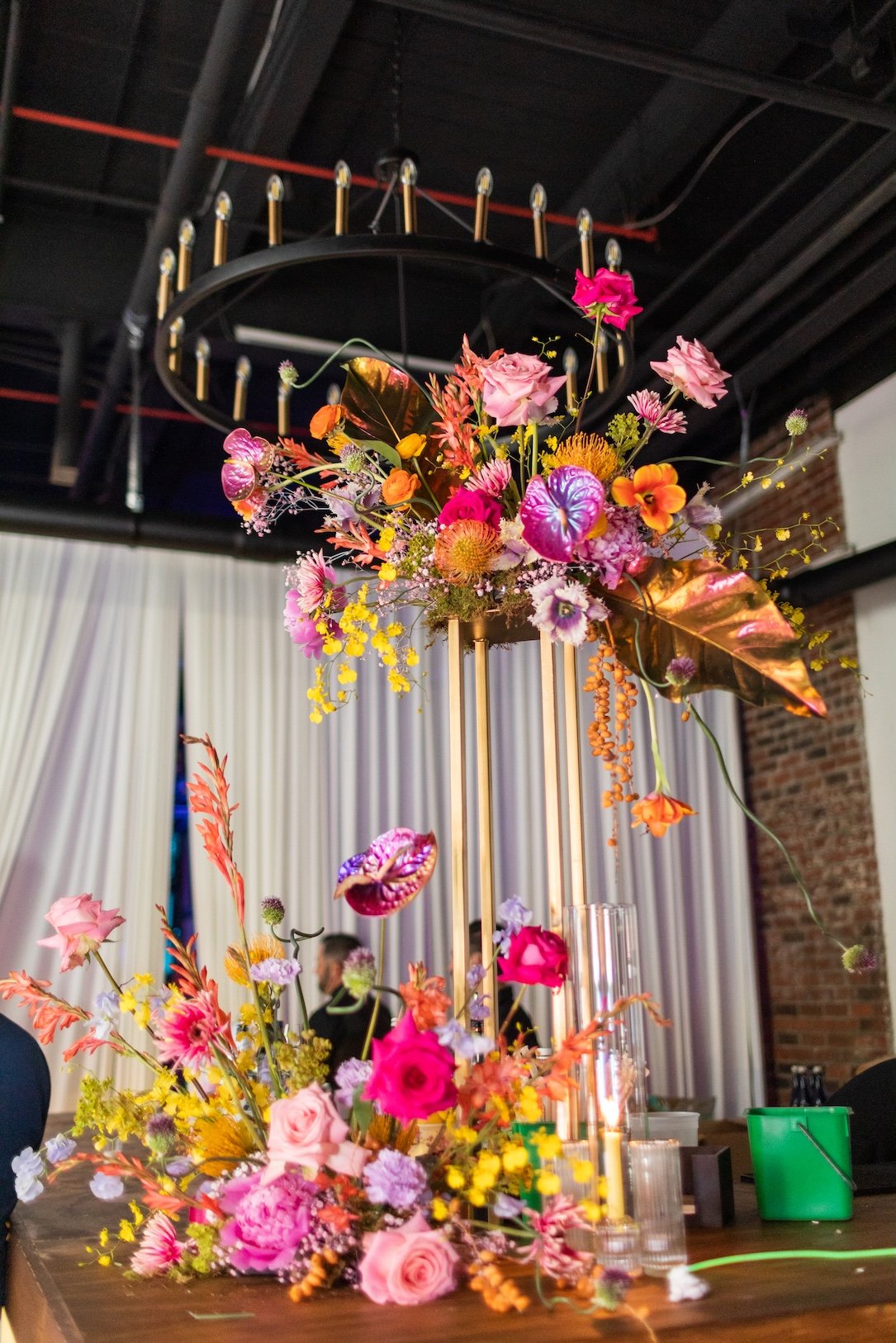 A vibrant floral centerpiece with various flowers in pink, purple, orange, and yellow, arranged on a table at an indoor event with a dark ceiling, exposed brick wall, and white curtains in the background.