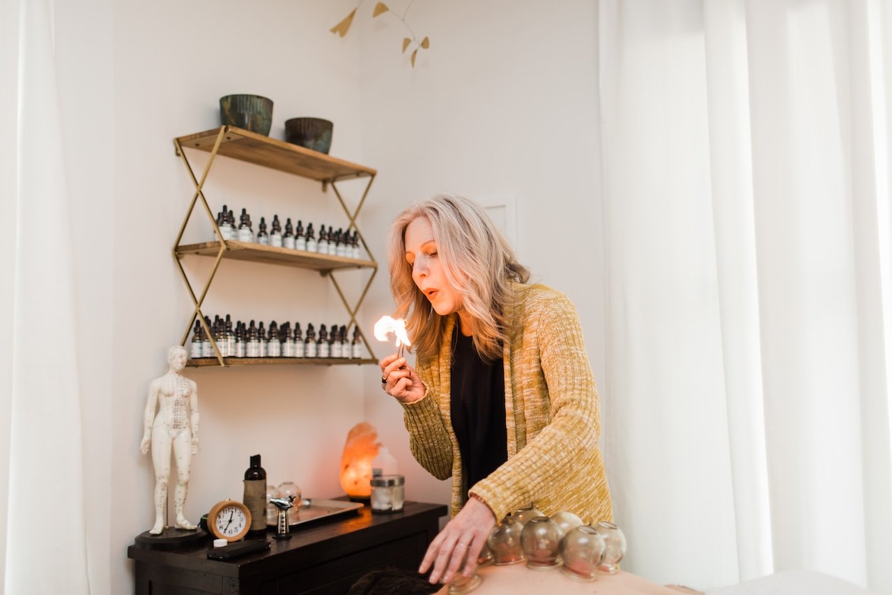 A woman with blonde hair wearing a yellow jacket, holding a burning object, performing a healing or ritual in a room with shelves holding essential oils, a lamp, and other wellness items.