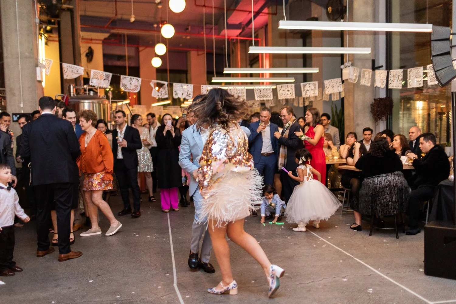 People dancing in a festive indoor event space with colorful lighting and decorative banners, surrounded by guests in formal and semi-formal attire.
