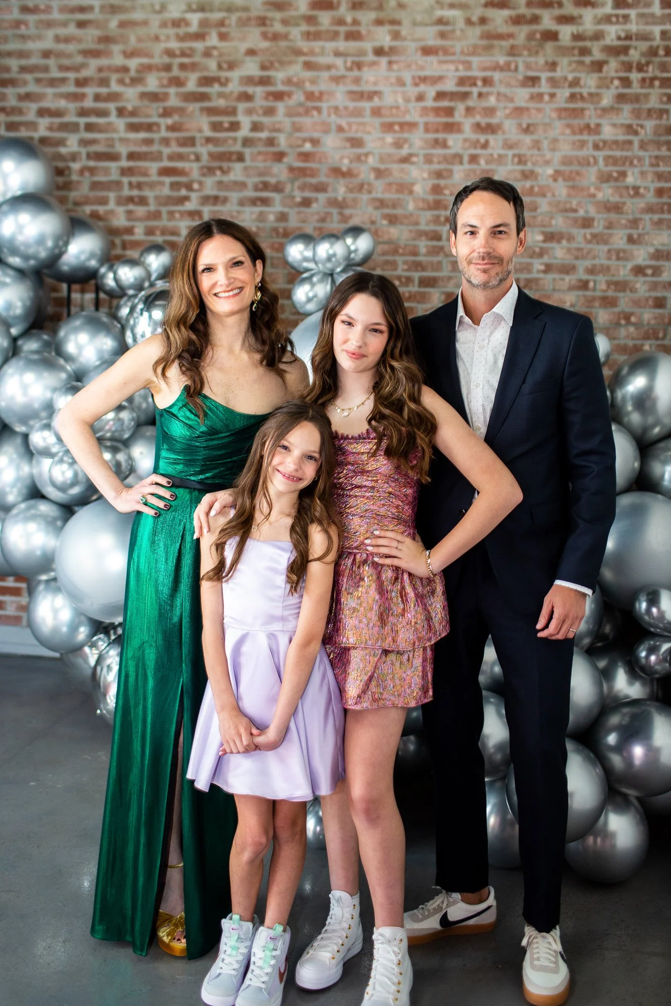 A family of five posing for a photo with balloon decorations in the background, standing in front of an exposed brick wall.