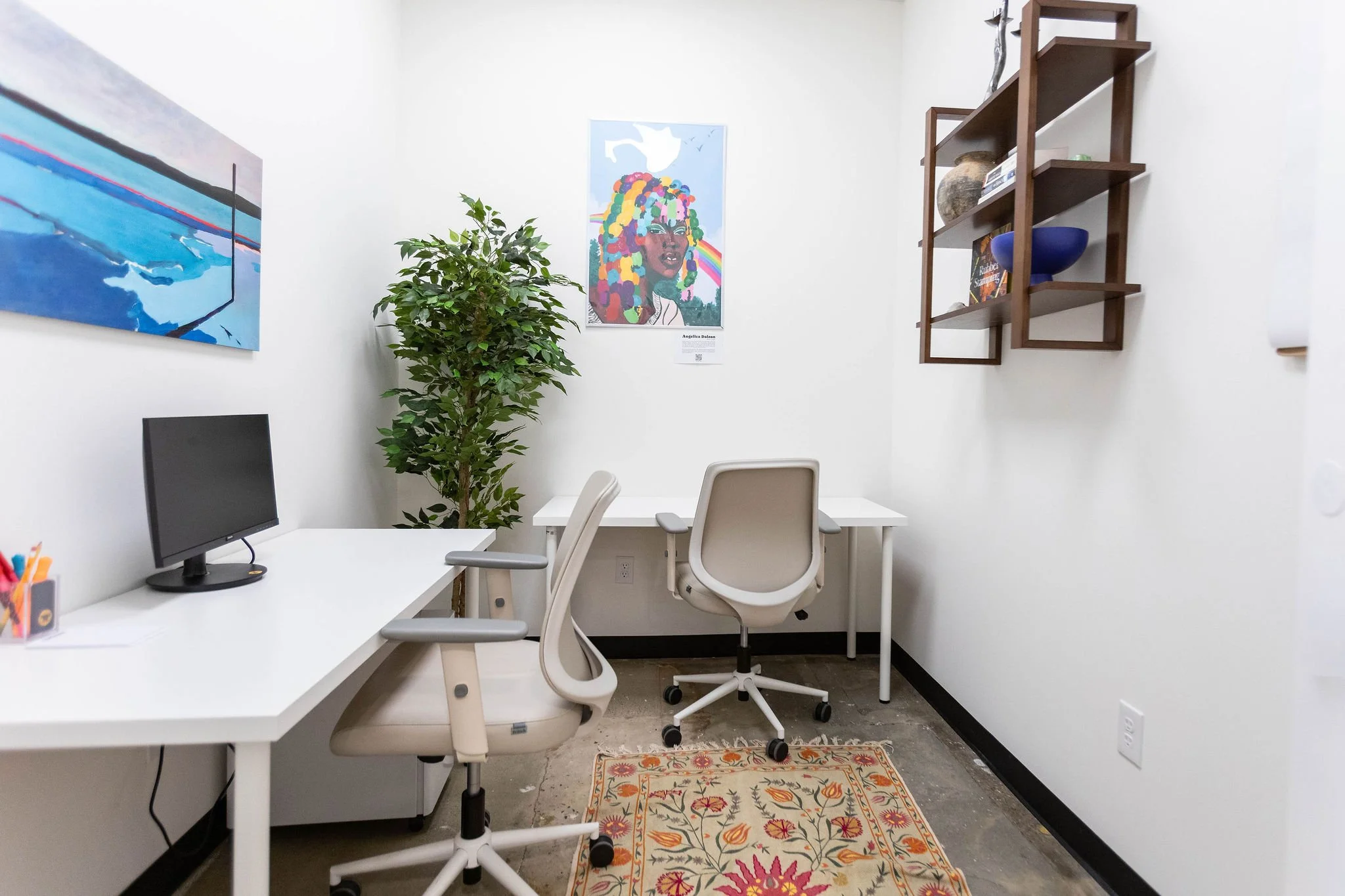 An office with two white desks and matching chairs, a computer monitor on one desk, a colorful rug, a green plant, and artwork on the walls, including a large colorful portrait of a woman with dreadlocks.