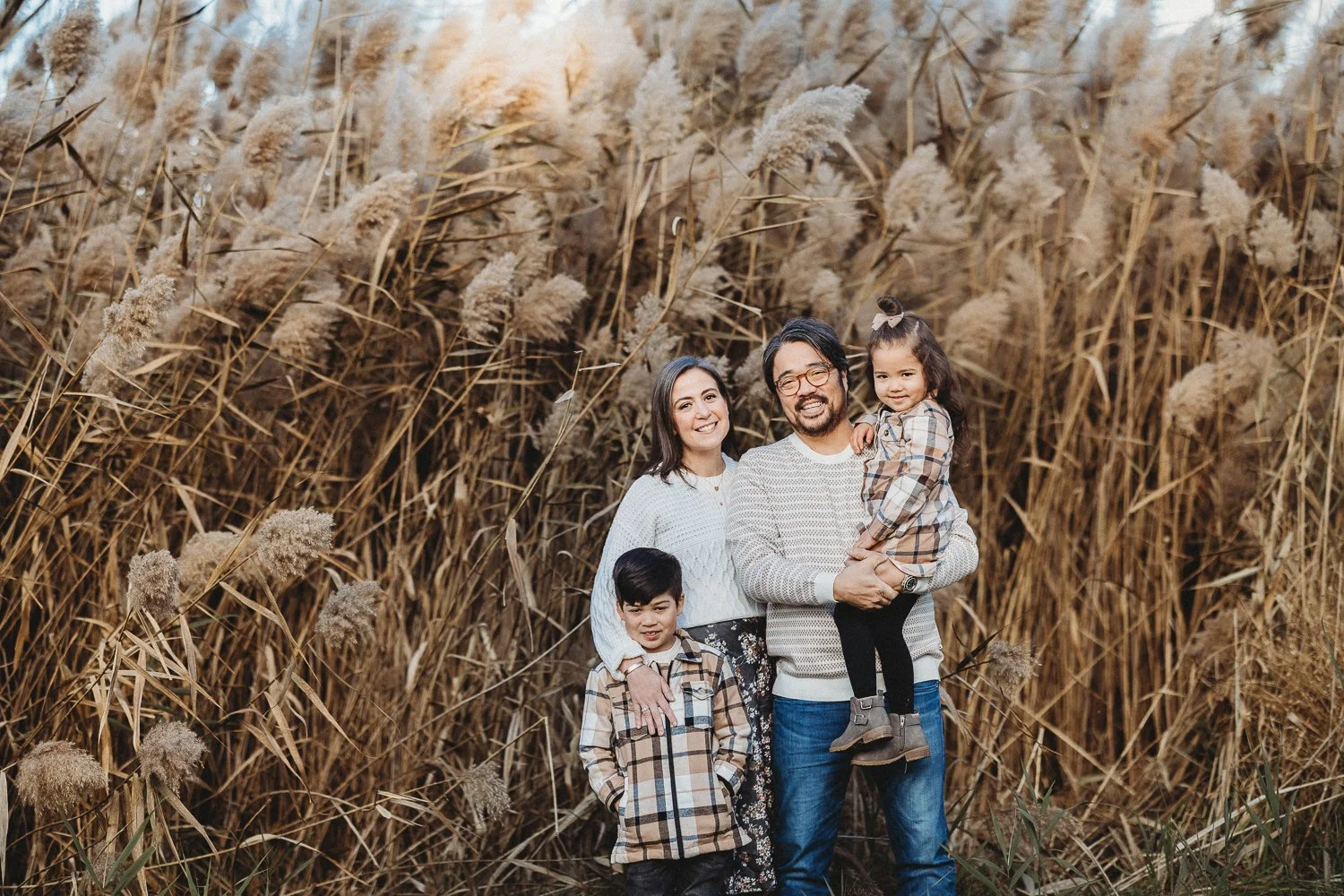 A young family of four posing in front of dry, tall grasses in Verona Park, NJ  for fall family mini session photos.