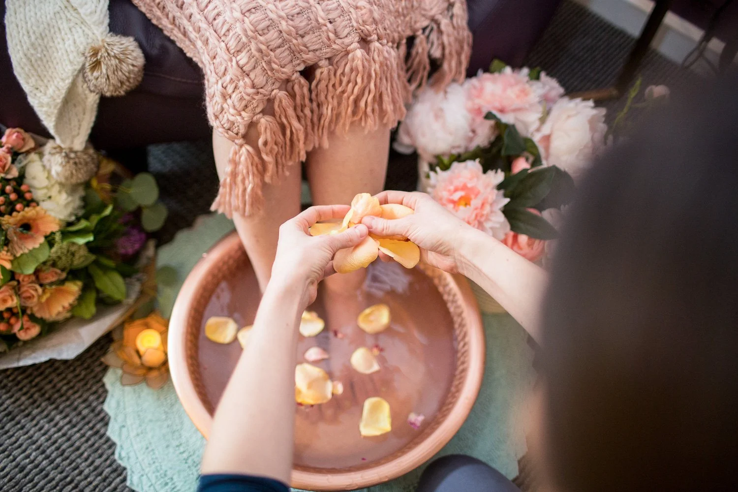 A person is giving a flower to another person who is sitting on a chair, surrounded by pink and orange flowers, with flower petals in a pink basin below.