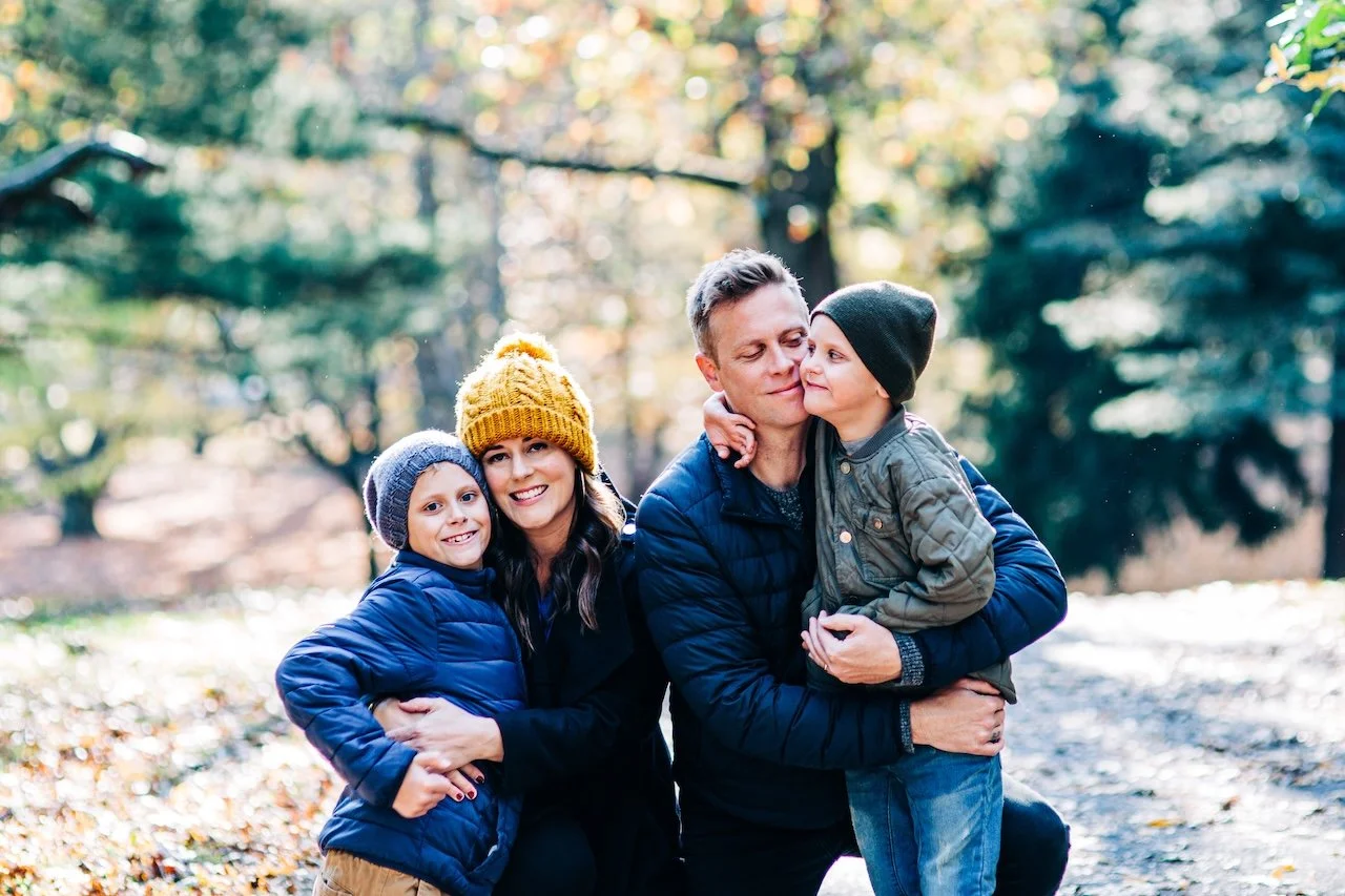 Family of four enjoying time outdoors in a forested park during autumn, wearing warm jackets and beanies.