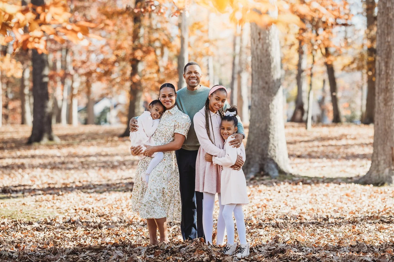 A Black family of five standing together in a park with autumn leaves on the ground and trees with orange and brown leaves, smiling for a photo.