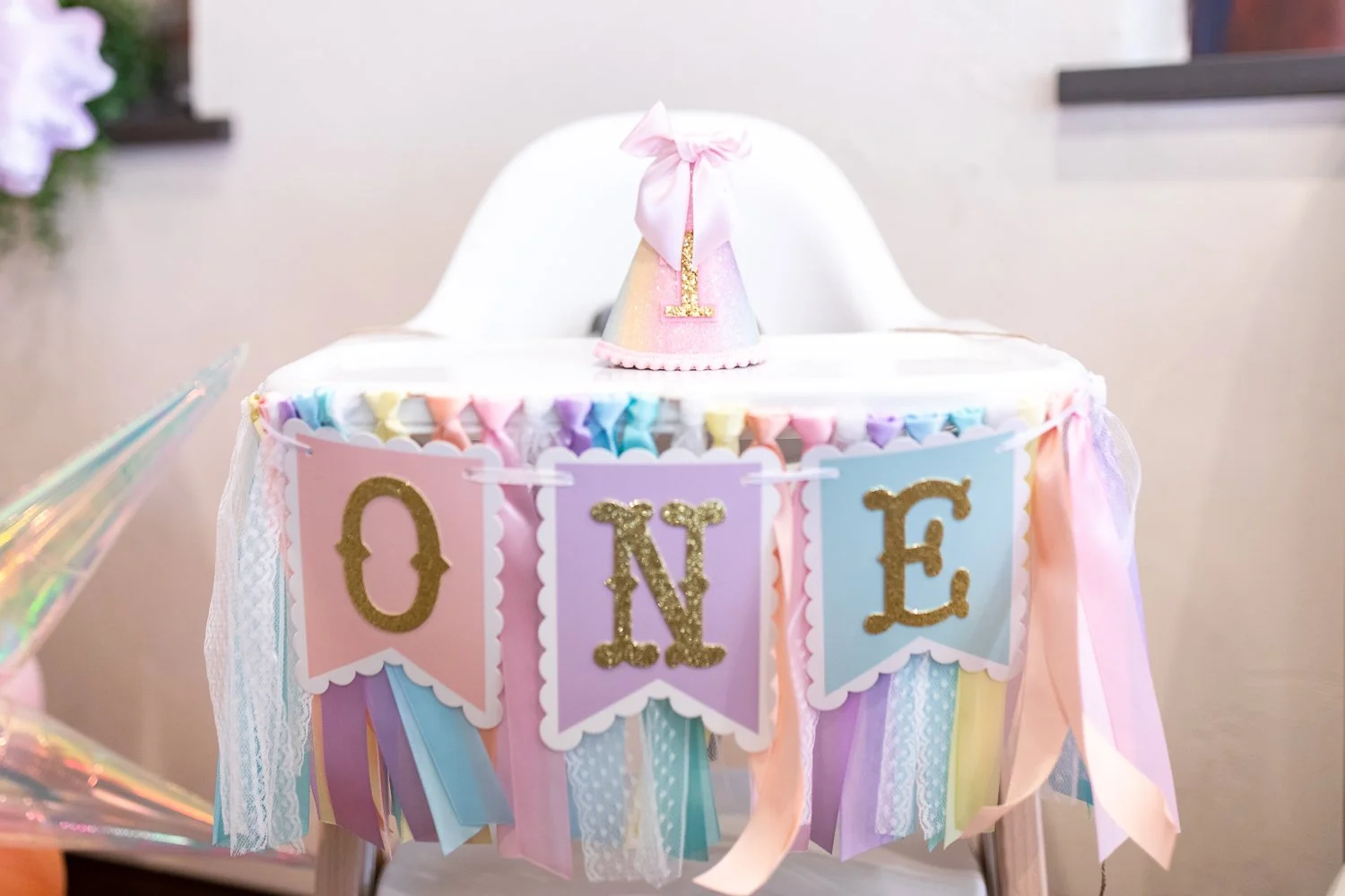 Colorful birthday cake with pastel-colored ribbons, a small pink and gold party hat with the number one, and a banner spelling 'ONE' in gold glitter letters.