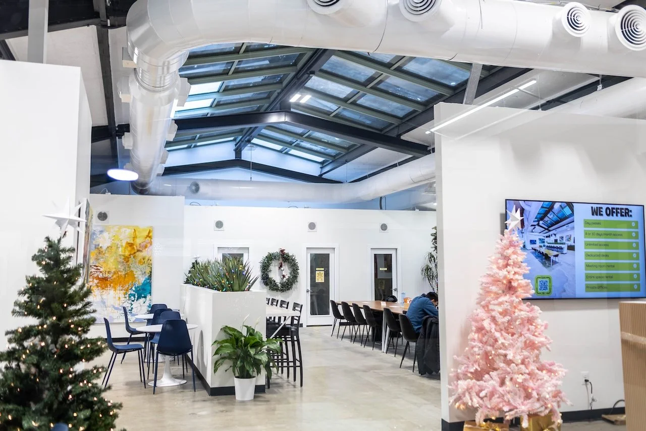 Modern indoor space decorated for Christmas with pink and green Christmas trees, a wreath on the door, and a colorful abstract painting on the wall. There are tables and chairs, a large digital display screen, and a glass ceiling allowing natural lig
