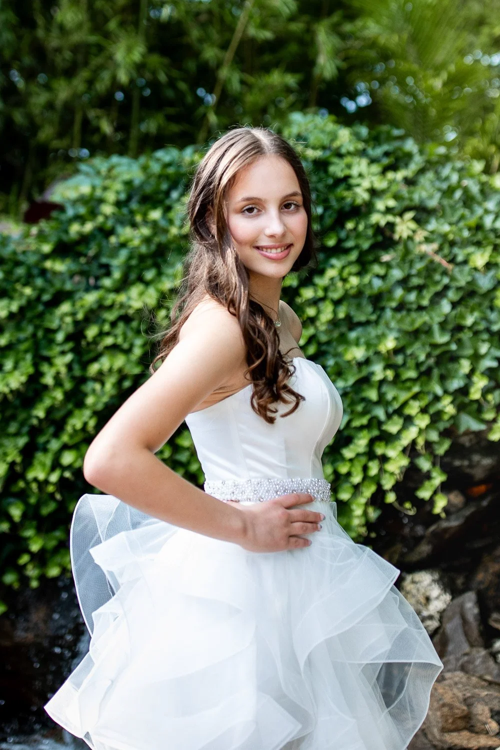 A young woman in a white strapless dress with a bejeweled waist stands outdoors with green foliage in the background, smiling at the camera.