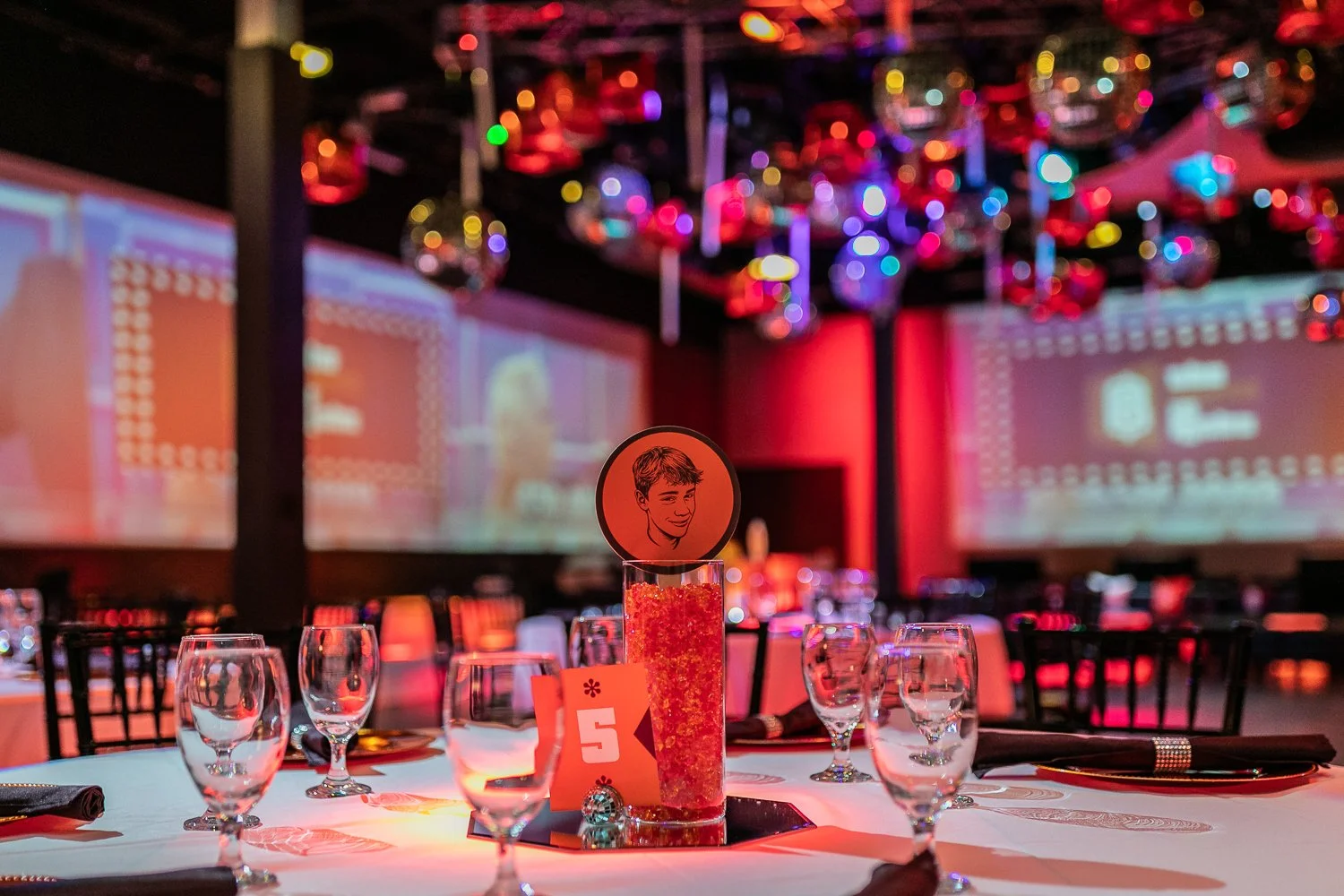 A decorated banquet table with glasses, black napkins, and a table number 5 card, in a ballroom with colorful disco balls hanging from the ceiling and large projection screens in the background.