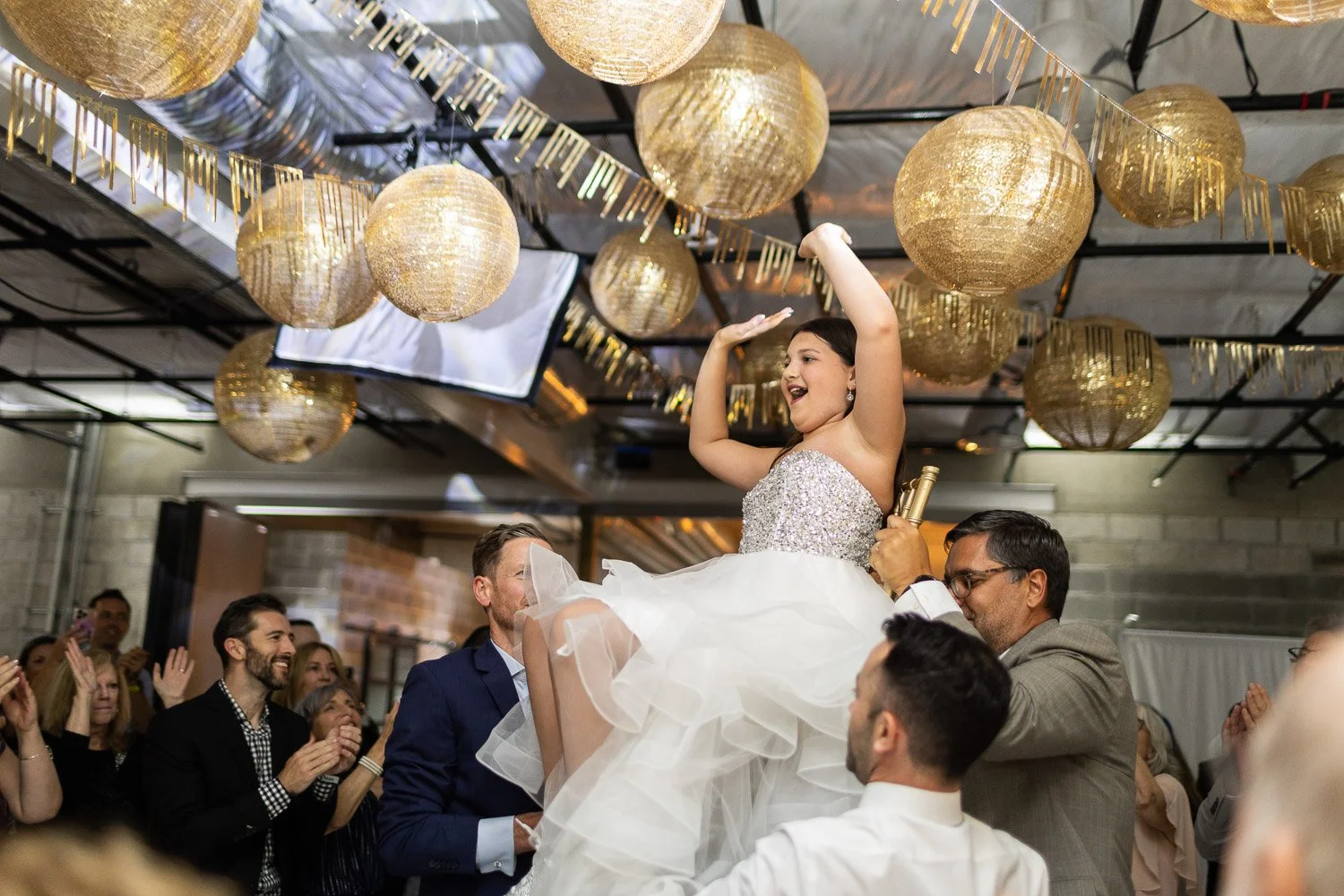 A Bat Mitzvah being lifted in a chair during a celebration at her party, surrounded by guests applauding and smiling, with decorative gold lanterns hanging from the ceiling.