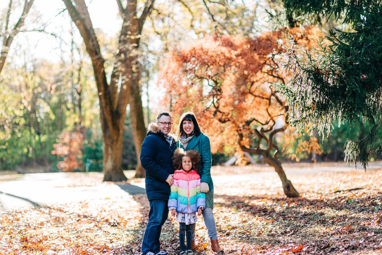 A family of three standing outdoors in autumn, smiling at the camera, with colorful trees and fallen leaves in the background.
