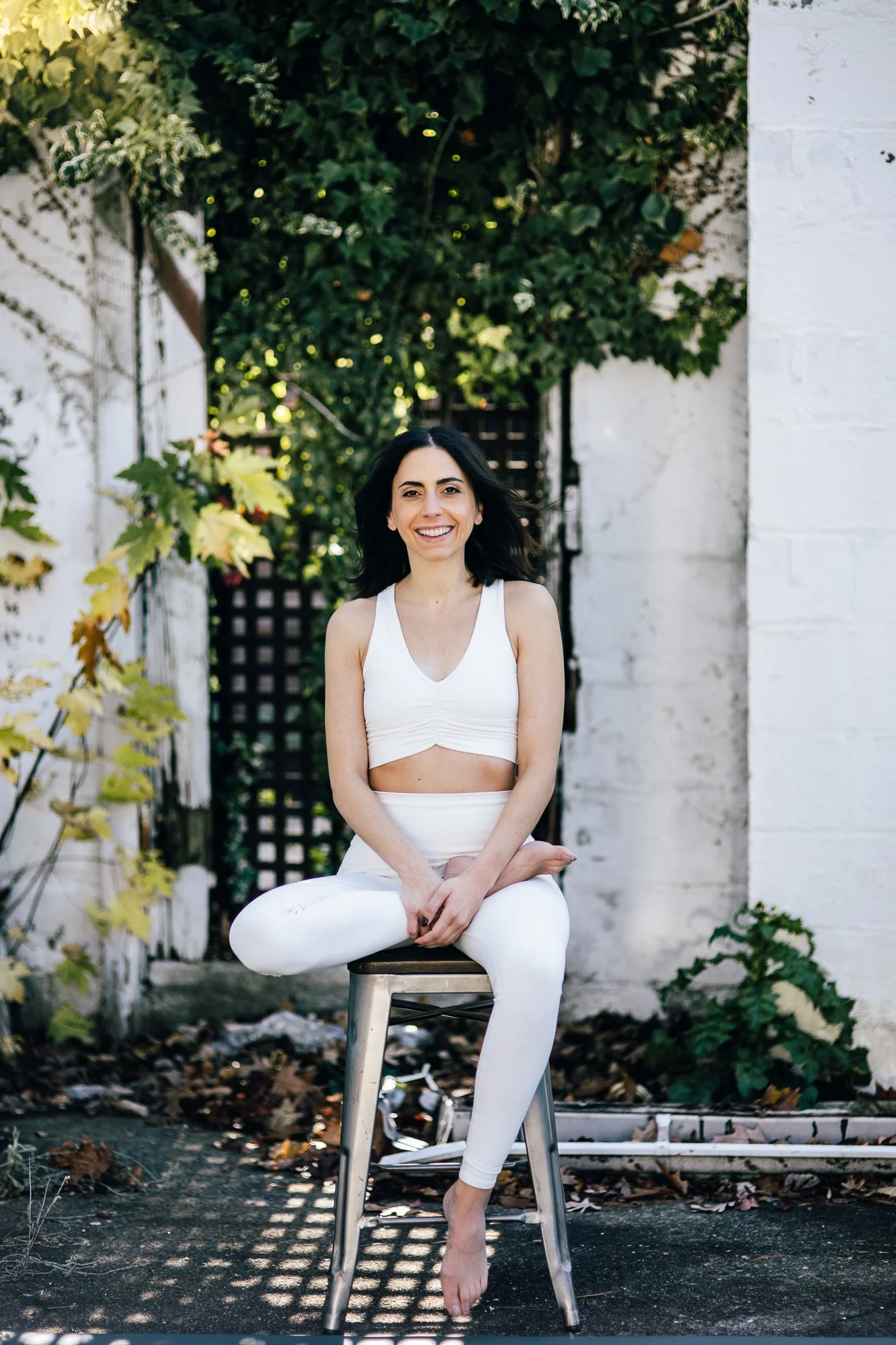 A young woman sitting cross-legged on a stool outdoors. She is smiling, wearing a white crop top and white leggings, with dark hair. The background includes a white wall, greenery, and trees with sunlight filtering through.