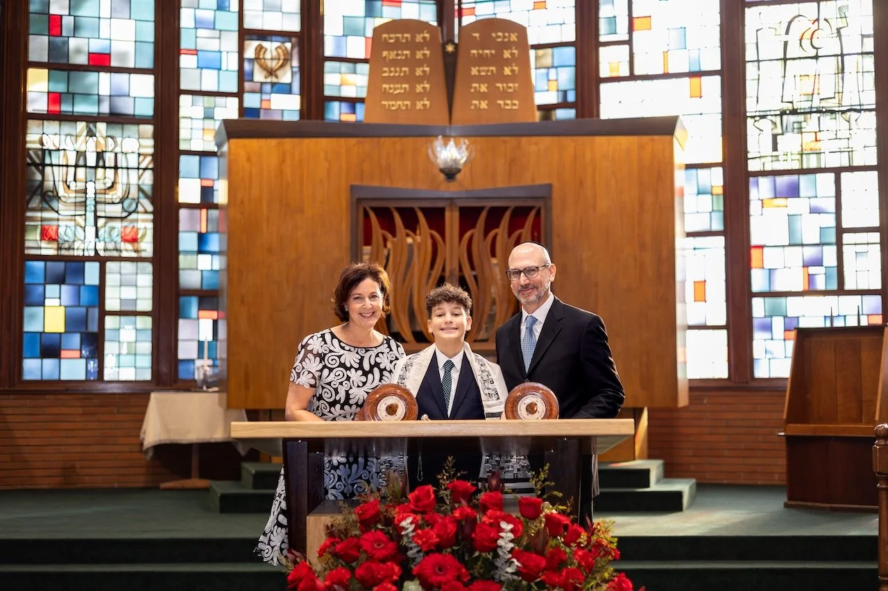 Three people, a woman, a boy, and a man, standing inside a synagogue in front of the Torah ark. The woman and man are smiling, and the boy is wearing a tallit. There are red flowers on the table in front of them, and stained glass windows in the back