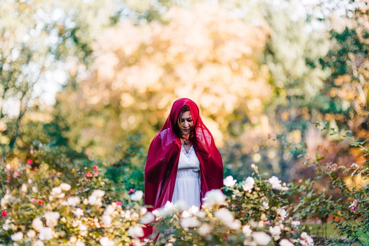 A woman in a white dress with a red shawl over her head, standing among white and pink flowers in a garden with autumn trees in the background.