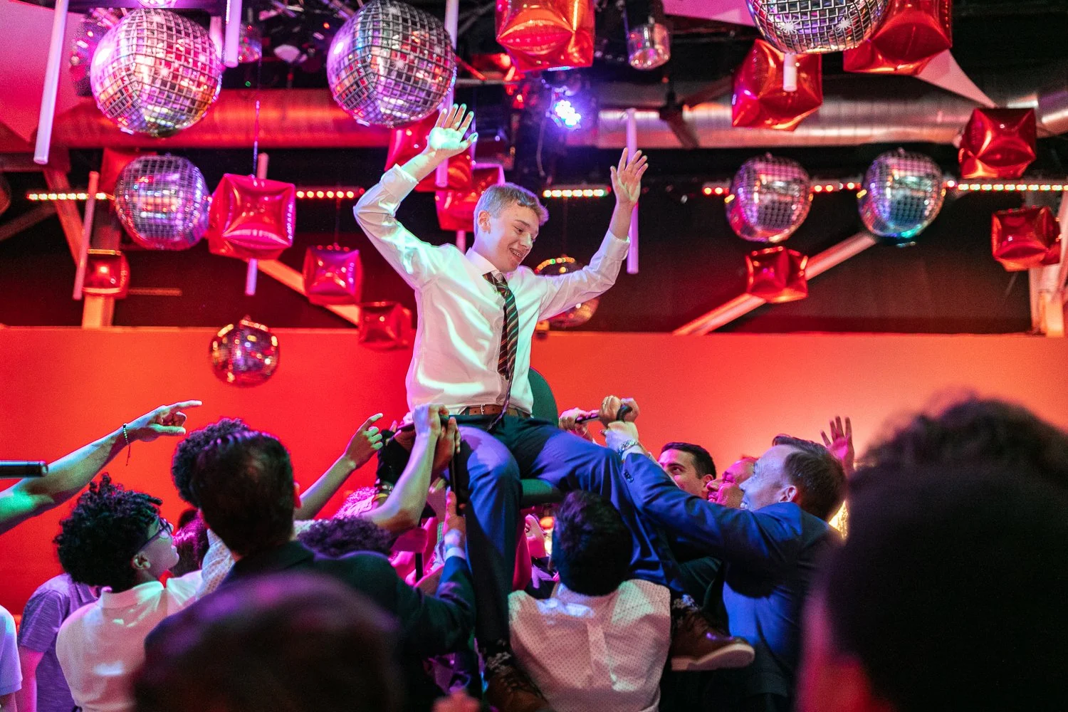 A young man wearing a white shirt and striped tie being lifted onto people's shoulders during a celebration in a party setting with disco balls and red star-shaped balloons hanging from the ceiling.