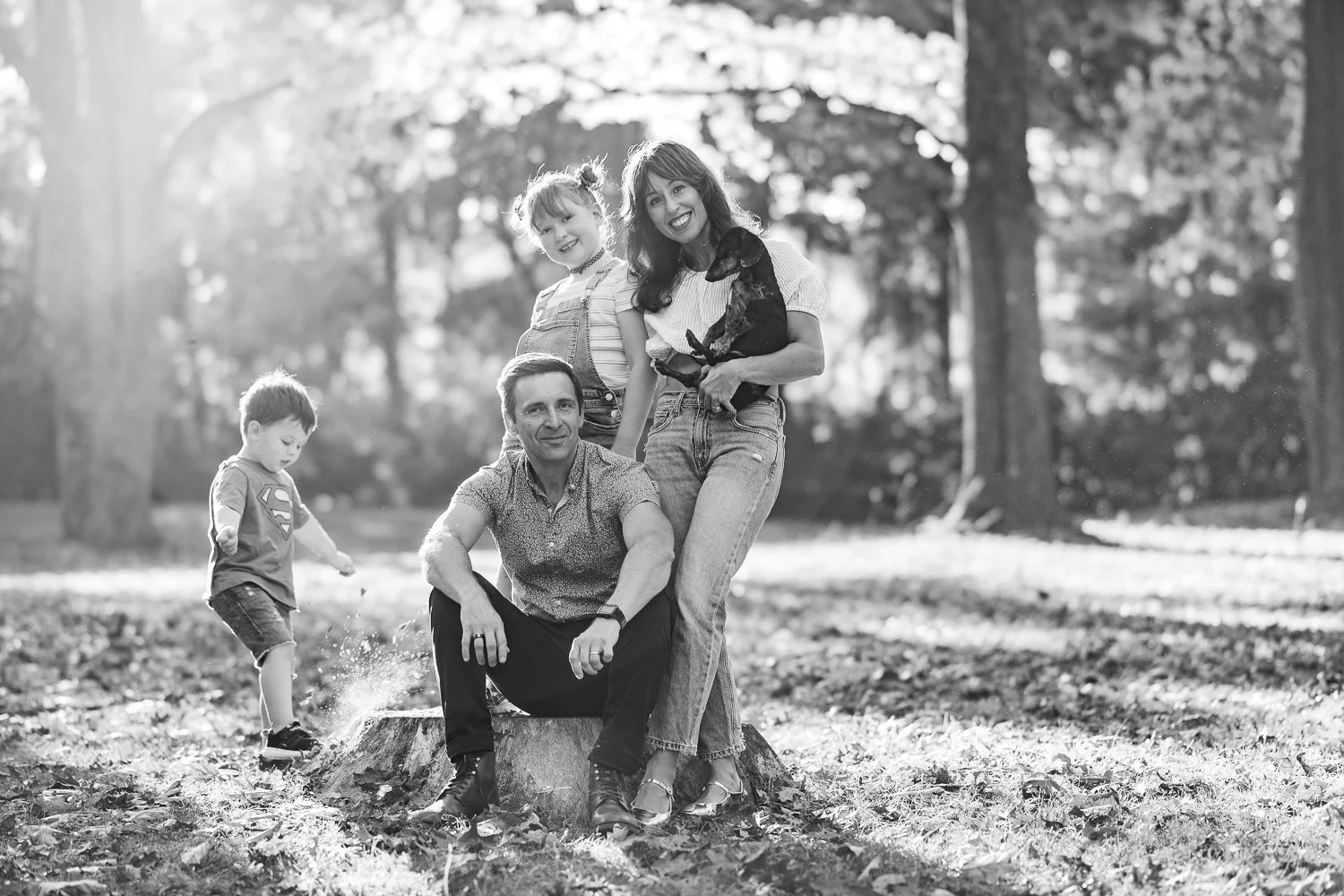 A family of five, including two children, an adult man, an adult woman, and a young boy, enjoying outdoor time in a park with trees and fallen leaves, black and white photograph.