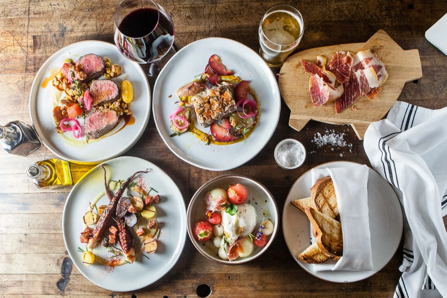 A spread of various dishes including sliced seared meat with vegetables, a piece of terrine with pickled onions, cured meats on a wooden board, grilled octopus with vegetables, a bowl of mozzarella and cherry tomatoes, and a basket of toasted bread, 