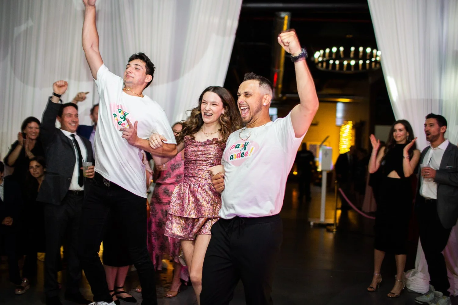 Bar Mitzvah girl entrance with happy people dancing and cheering at an elegant bat mitzvah, some wearing white T-shirts with colorful text, others in formal attire, with a festive atmosphere and smiles.