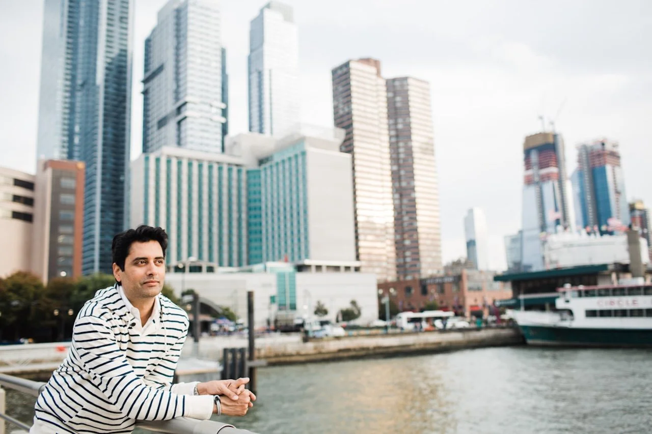 A man leaning on a railing along a waterfront in front of tall city skyscrapers and a boat docked nearby.
