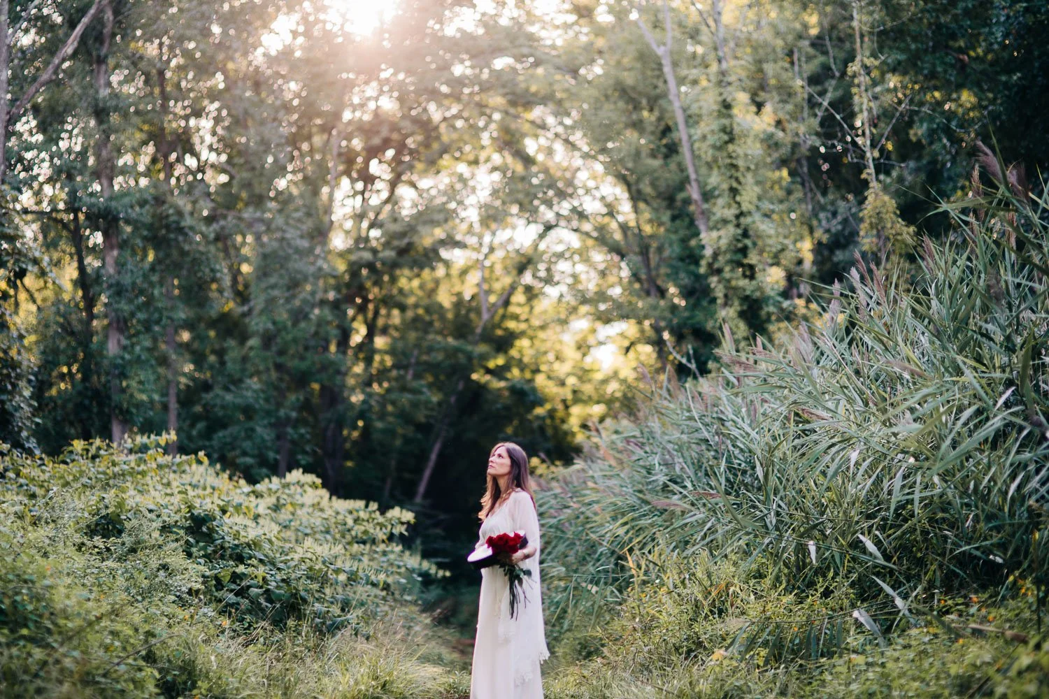 A woman in a white dress holding a bouquet of red flowers in a lush, green forest during sunset.