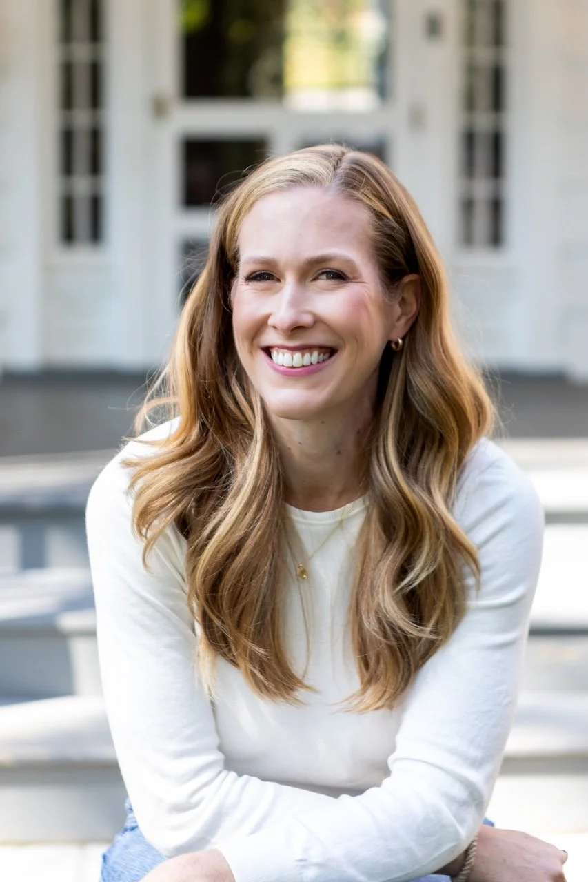 A woman with long, wavy, light brown hair smiling outdoors, wearing a white top and small gold hoop earrings, sitting on steps in front of a white building with windows.