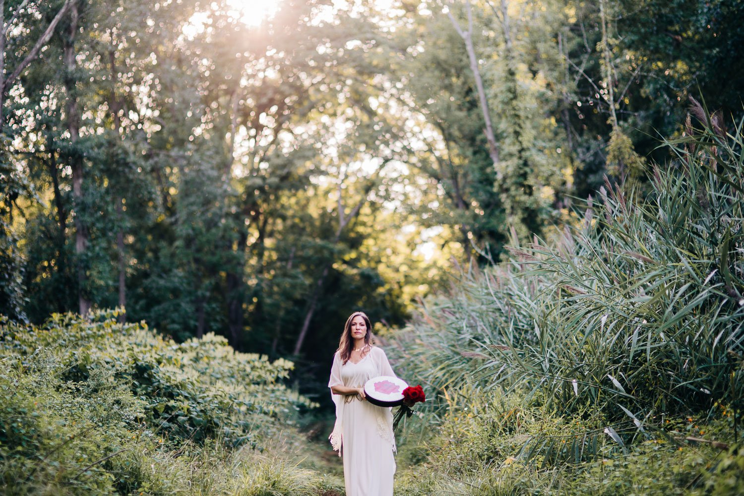 A woman in a long white dress holding a round object with pink and white design and a bouquet of red flowers, standing on a grass path surrounded by tall green plants and trees, with sunlight shining through the trees.