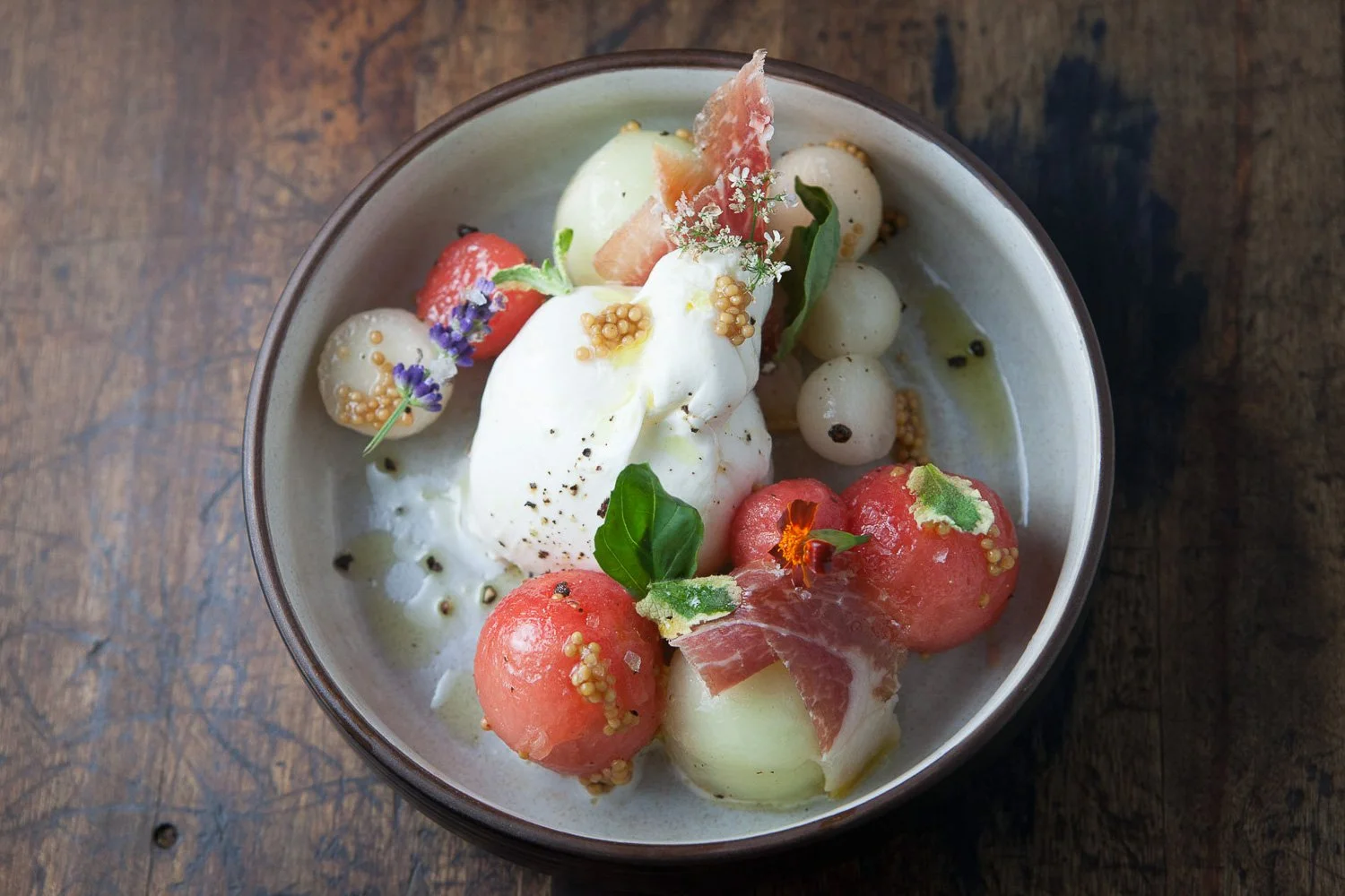 A bowl of melon balls, strawberries, whipped cream, and edible flowers garnished with herbs and mustard seeds.