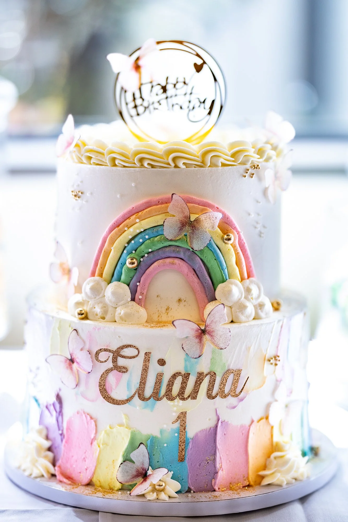 A colorful birthday cake decorated with pastel rainbow-colored fondant arcs, white frosting, edible butterflies, and a gold birthday topper. The cake has a white base with gold lettering that reads 'Aeliana' and a gold number one.