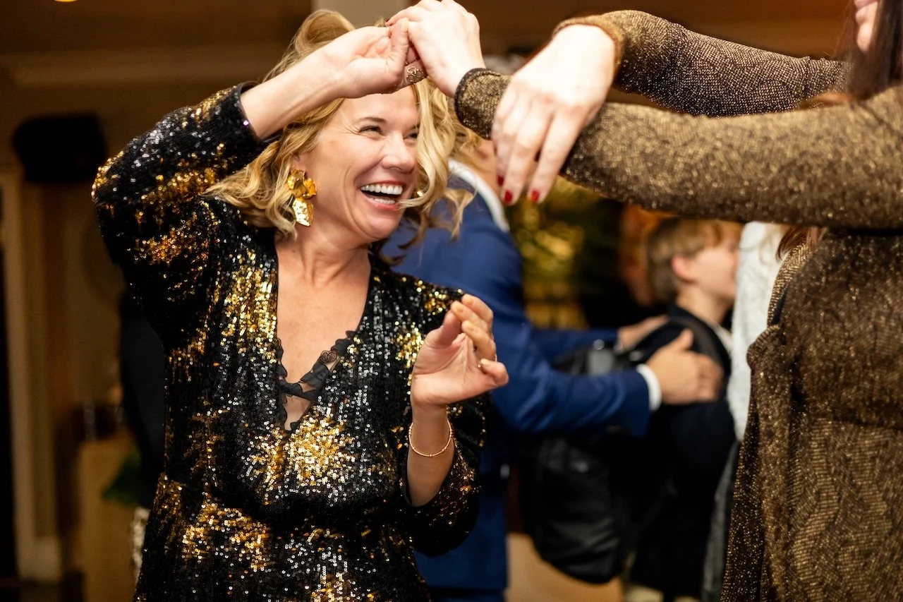 A woman with blonde hair wearing a black and gold sequin dress is dancing and laughing with another person at a party.