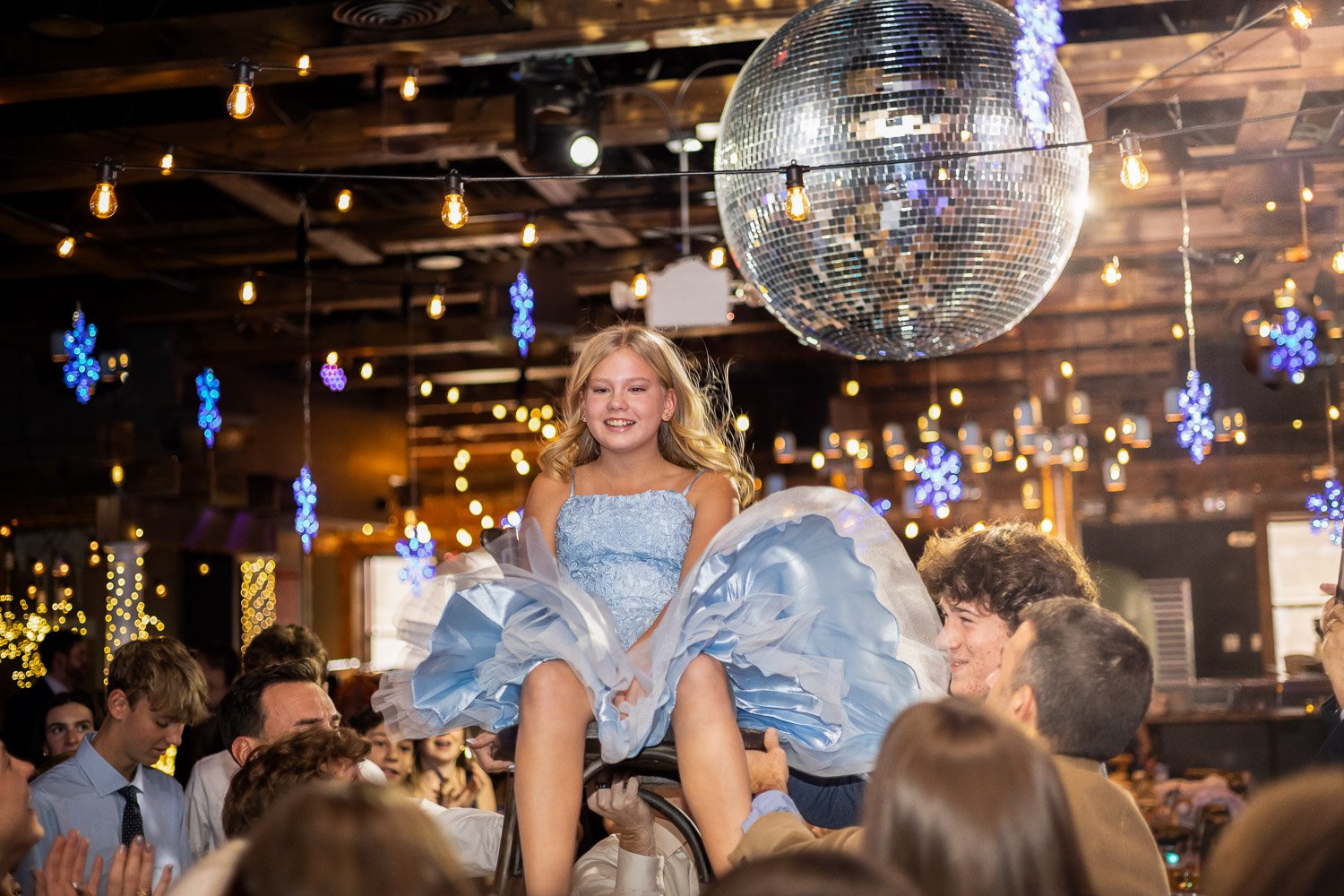 A girl in a blue dress being lifted on a chair by people at a Bar Mitzvah, with a large disco ball and Christmas lights in the background.