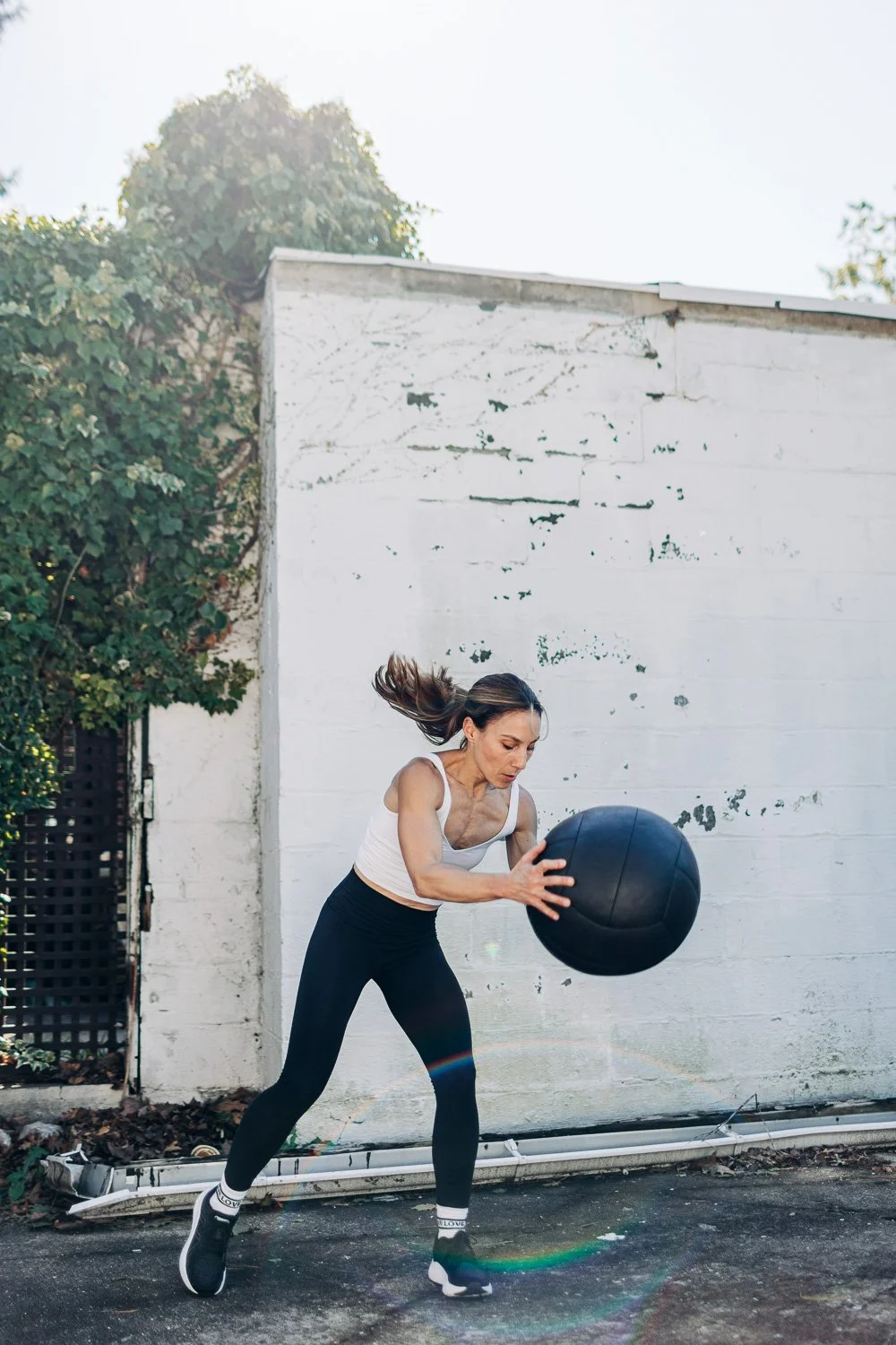 Woman in athletic clothing training outdoors with a medicine ball against a white wall, with greenery and bright sunlight in the background.