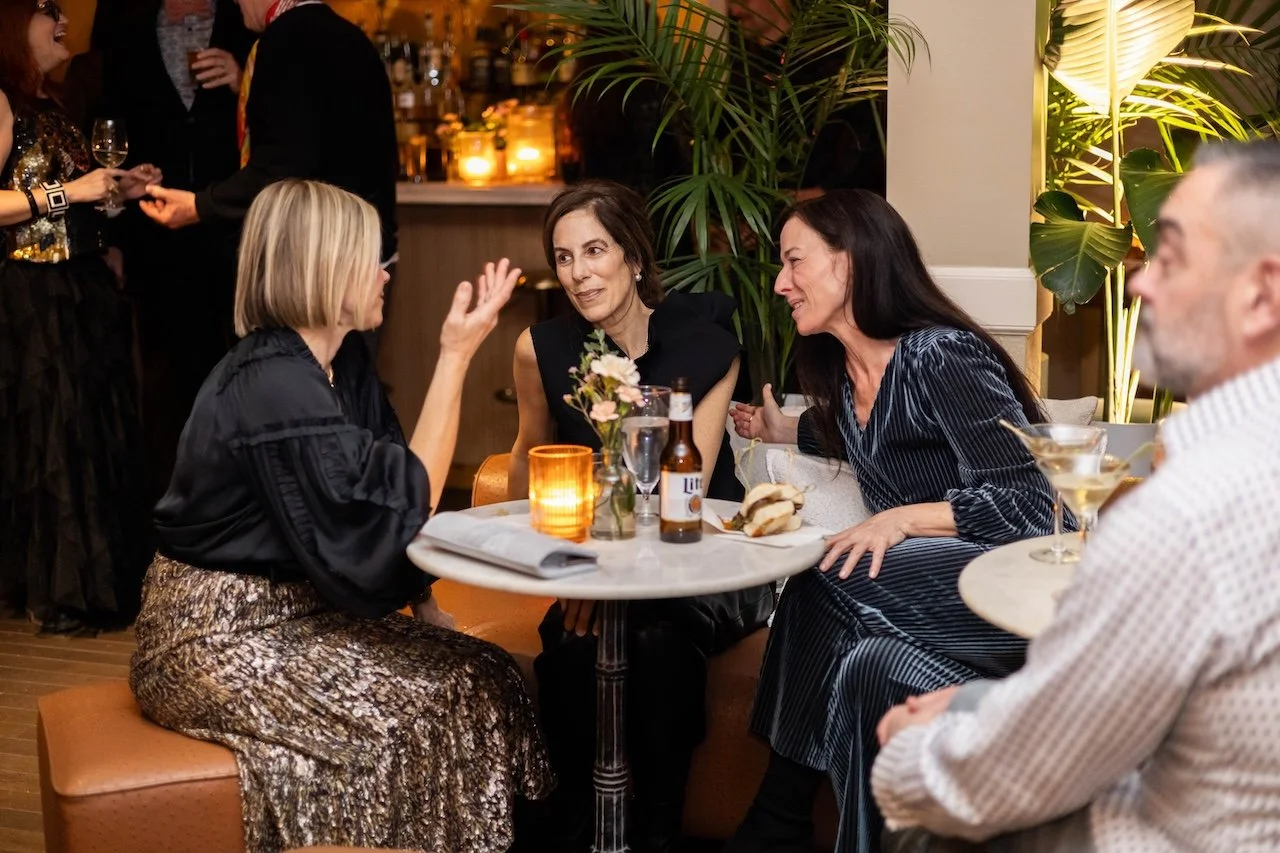 Four adults sitting at a round table having a lively conversation at a social gathering, with drinks and a floral centerpiece on the table, in a warmly lit indoor setting.