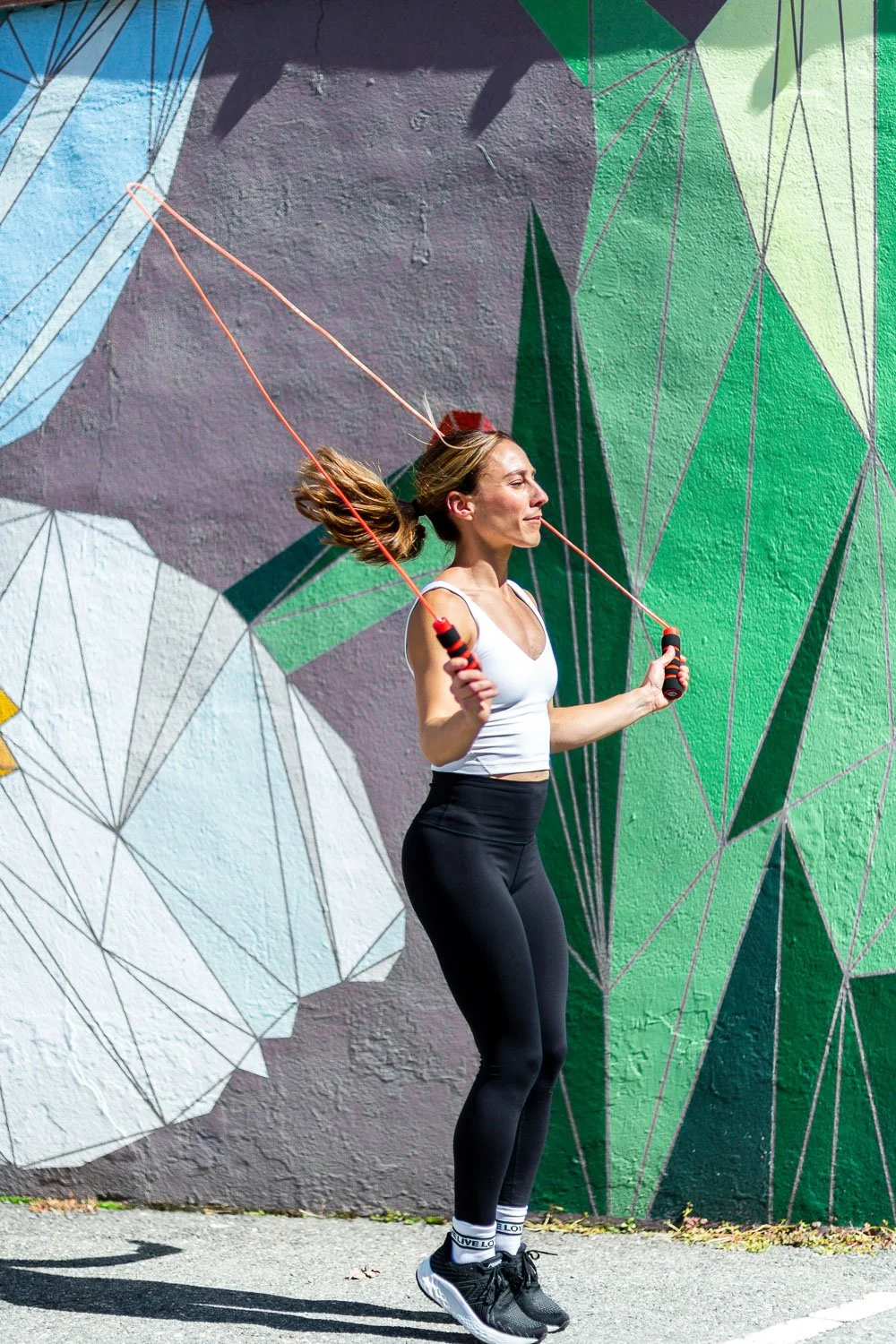 A woman skipping rope outdoors in front of a colorful geometric mural with shades of green, purple, and white.