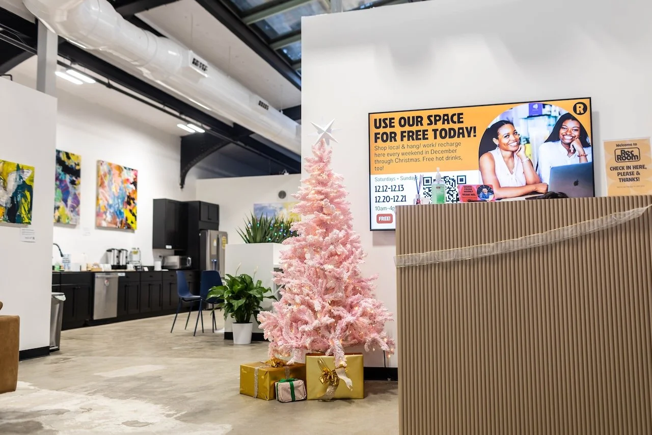 Pink artificial Christmas tree with gold and striped wrapped presents underneath, in a modern office space with abstract colorful artwork on the wall, a gray countertop with coffee makers, and a digital display promoting event space rental.